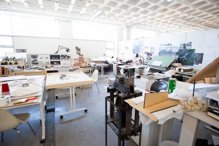 A brightly lit room in the John A. Russell Building for Architecture has many stacks of books and art supplies used to create models on the tables.