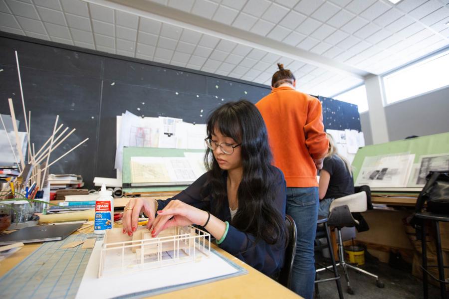 A student with glasses and long dark hair is seated at a table using wood and glue to build a model in a classroom in the John A. Russell Building.