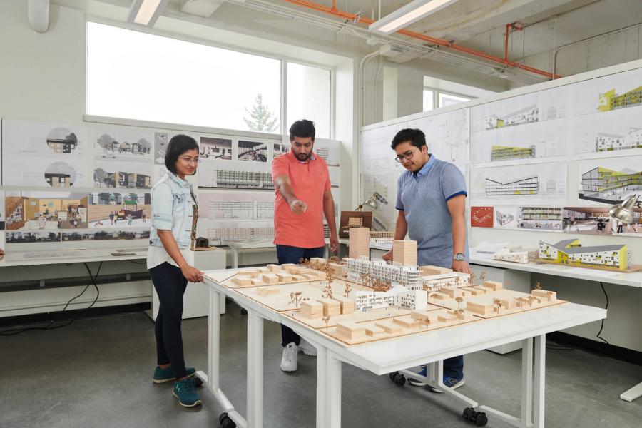 Three students stand in a John A. Russell Building architecture classroom looking at a small wooden model of a city.