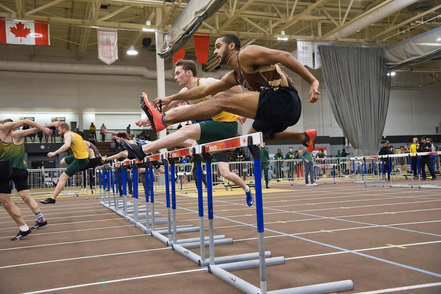 Two athletes in UM Bisons jerseys jump hurdles at the exact same time at a track and field event at the Max Bell Centre.