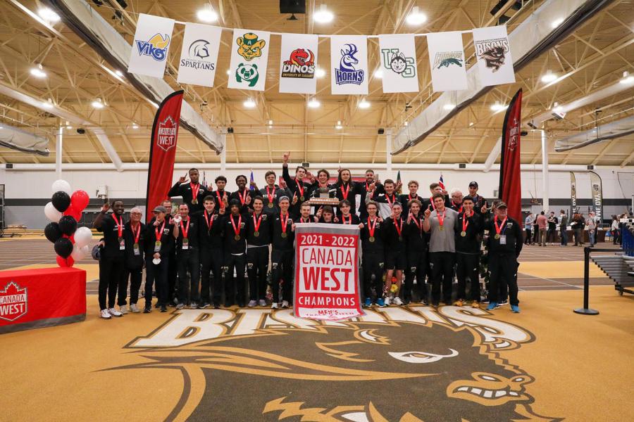 A large group of University of Manitoba Bisons athletes stand together in the gym of the Max Bell Centre wearing medals with red ribbons behind a sign that says 2021 - 2022 Canada West Champions Men's Track and Field.