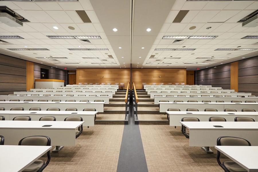 A Robson Hall Faculty of Law lecture hall with many rows of beige chairs at white tables from the perspective of a speaker at the bottom of the stairs at the end of the room.