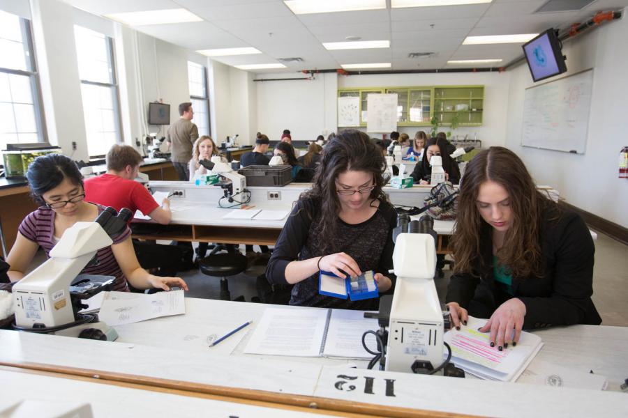 Students seated in a white Science Complex classroom at tables with microscopes, books, and papers.