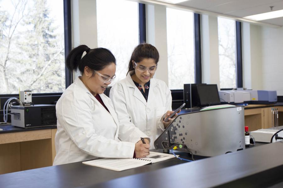 Two students wearing white lab coats and clear googles smile and look down at a book they are writing in on the table of a Science Complex classroom. There is a metal machine in front of them.