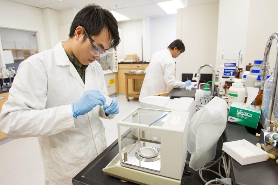 Two students in googles, white lab coats, and blue gloves test samples and write down data in a laboratory.