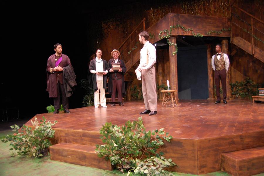 Five actors standing on stage at the Taché Arts Complex. They are wearing costumes from the early 1900s and a brown stage is decorated with scattered green fake plants.