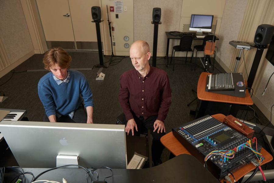 A student and an instructor seated in a small studio room in the Taché Arts Complex in front of an Apple computer monitor. There are speakers around the room and mixing boards with small switches and knobs on the table.