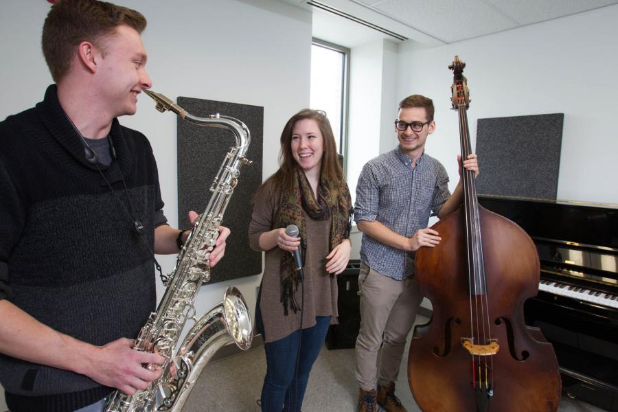 Three students stand laughing and talking in the Taché Arts Complex, one holding a cello, one holding a saxophone, and one holding a microphone.