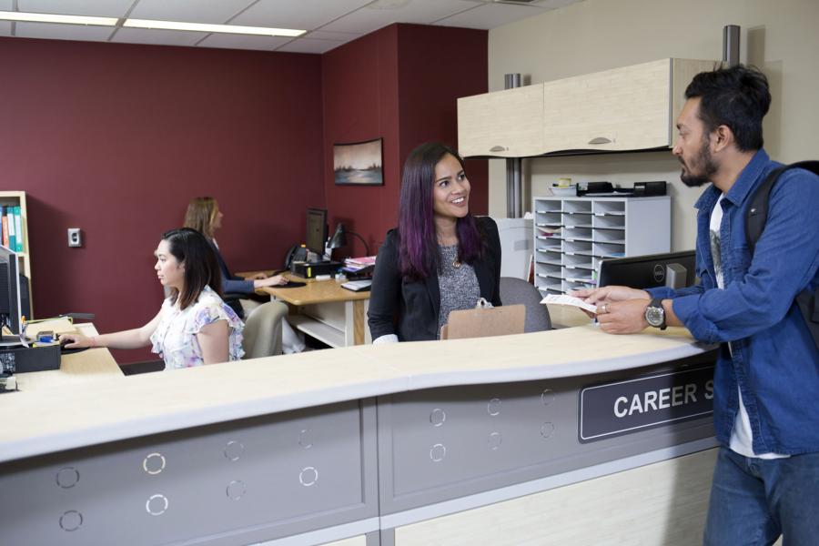 Three people work behind the Career Services desk at the UMSU University Centre as a student in a long sleeve blue shirt stands on the other side of the desk looking for help.