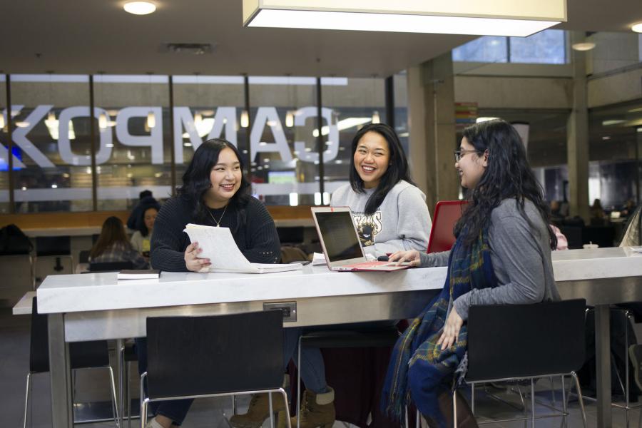 Three students with long hair sit smiling and talking at a white table in the Campo area of the UMSU University Centre building.