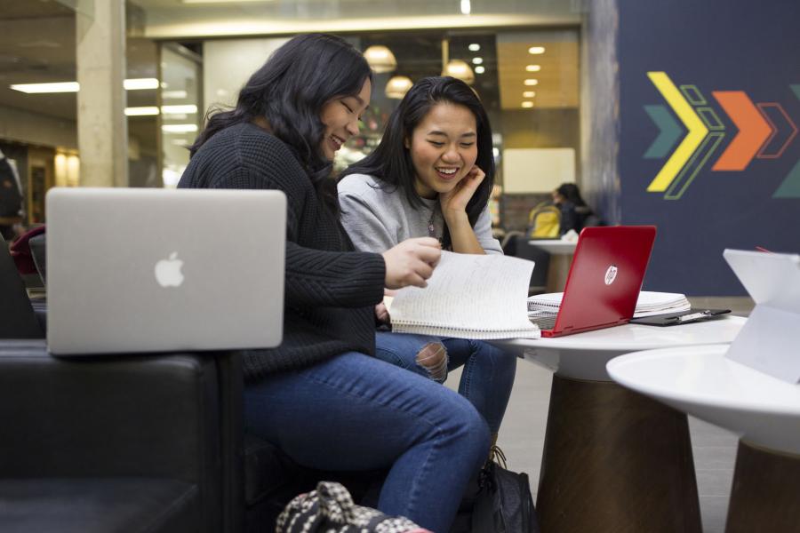 Two students sit with notebooks and laptops in the main area of the UMSU University Centre building.