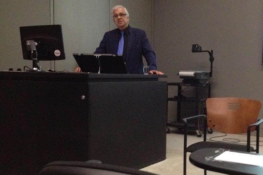 Robert Houle stands behind a lecture plinth, facing the camera, in the middle of presenting.