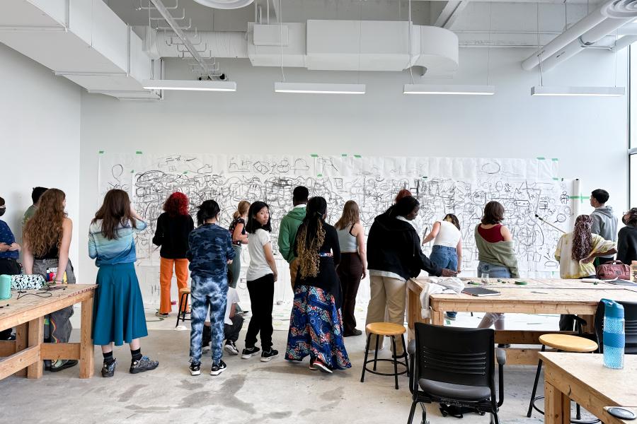A group of students collaboratively sketch on a large sheet of paper taped to a studio wall.