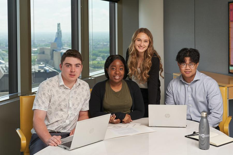 Four Asper business students sitting at a board room table with downtown Winnipeg in the background.