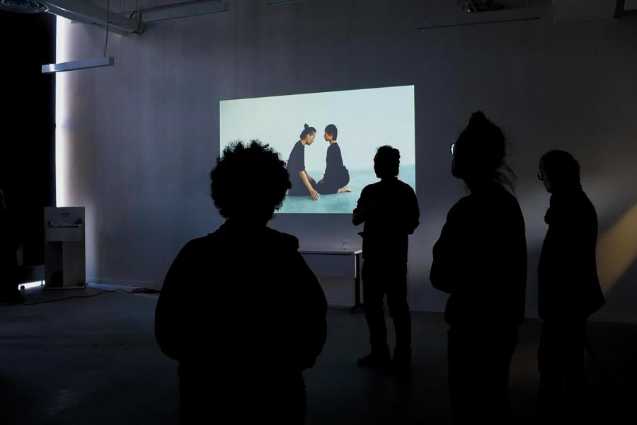 Viewers watch a video installation of two figures sitting face-to-face on a gallery wall.