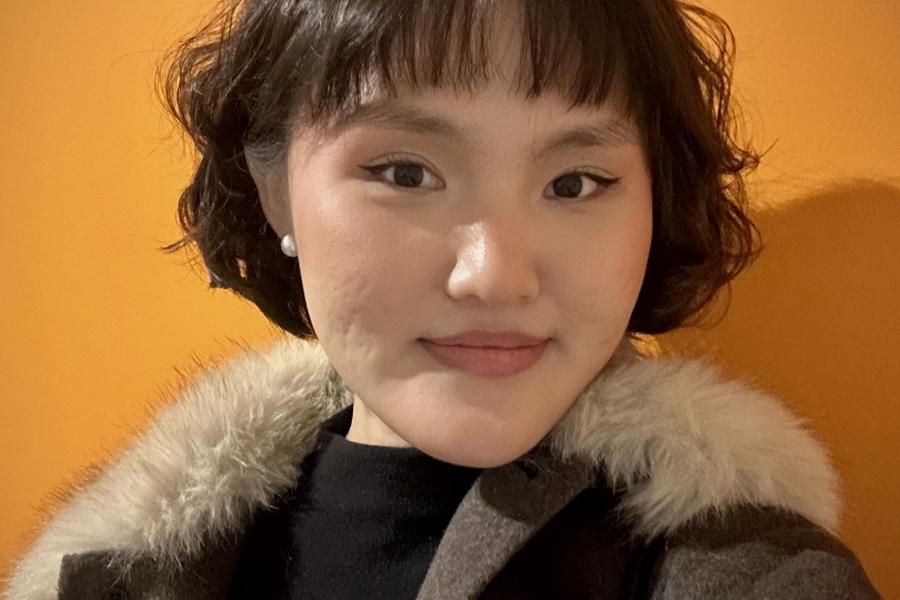 Portrait of Alyssa Kim with short wavy hair, wearing a fur-trimmed coat, standing against a bright orange wall.