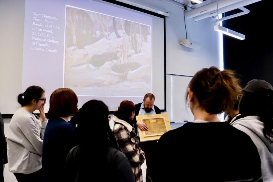 Professor showing a group of students the back of a painting, with a painting of a wintery nature scene displayed on the screen behind him.