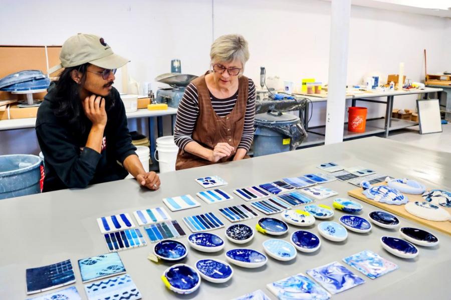A student with a professor who is reviewing their pottery work. Looks like a variety of shapes including dishes and coasters or tiles. 