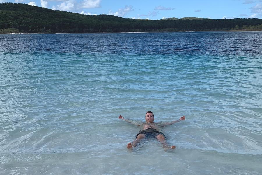 A student relaxing with arms outstretched in shallow water with mountains in the background