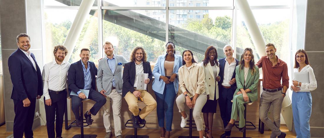 A diverse group, dressed in business casual attire, smiling and posing confidently in an office with large windows and natural light.