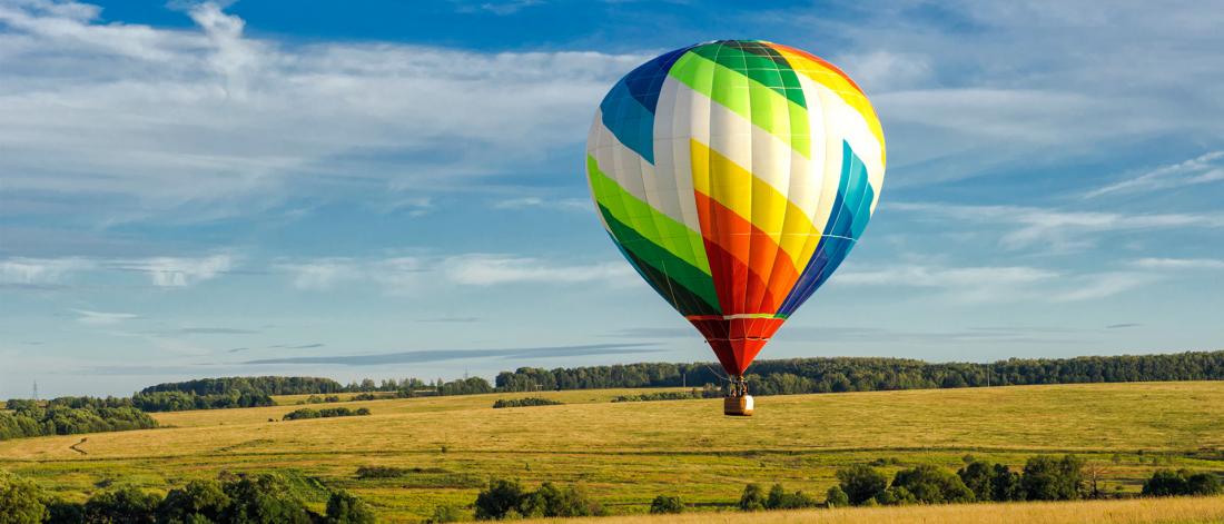 A colourful hot air balloon with rainbow stripes floats over a vast, sunlit meadow under a clear blue sky.