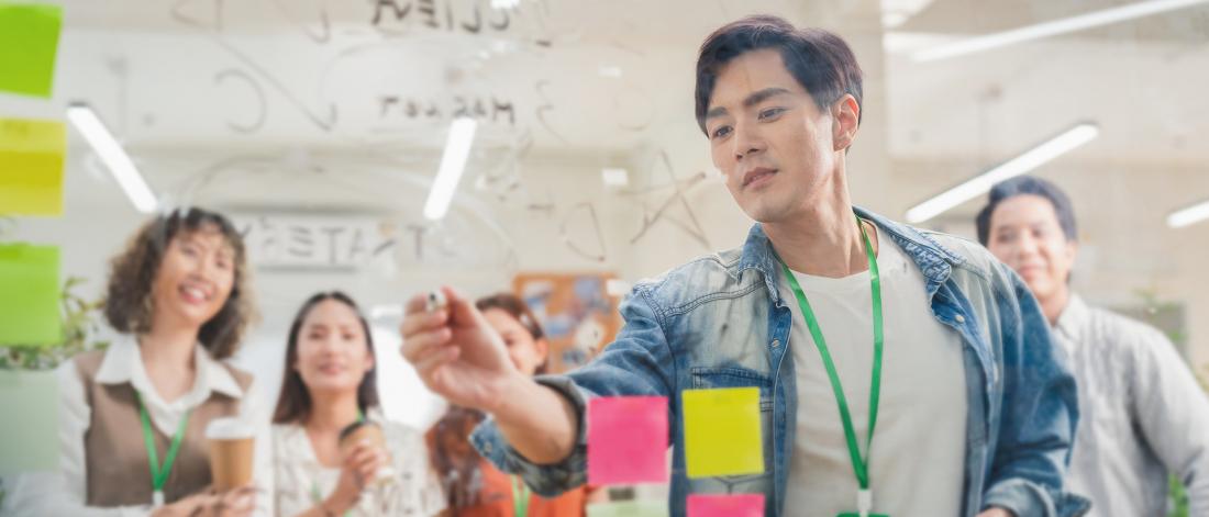 A group brainstorming session with a man writing on a transparent board covered in diagrams and notes while three colleagues watch attentively. 