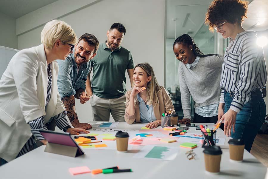 A diverse group of colleagues is collaborating around a table covered with colorful sticky notes, markers, and coffee cups, suggesting a brainstorming or planning session. 