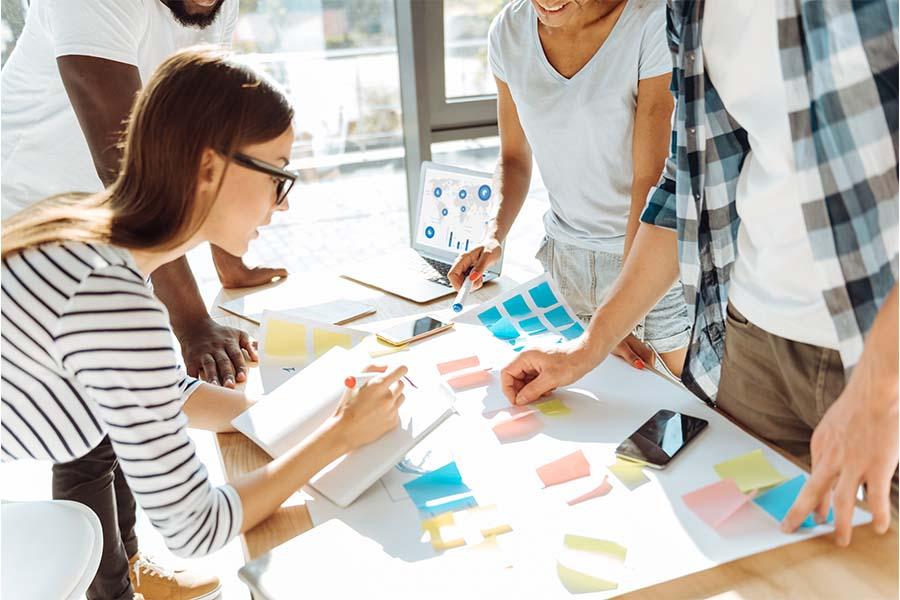 A group of people collaborating around a table covered with colorful sticky notes and papers, with a laptop displaying charts in the background. The scene highlights teamwork and brainstorming, emphasizing idea organization and planning through visual aids and active discussion.