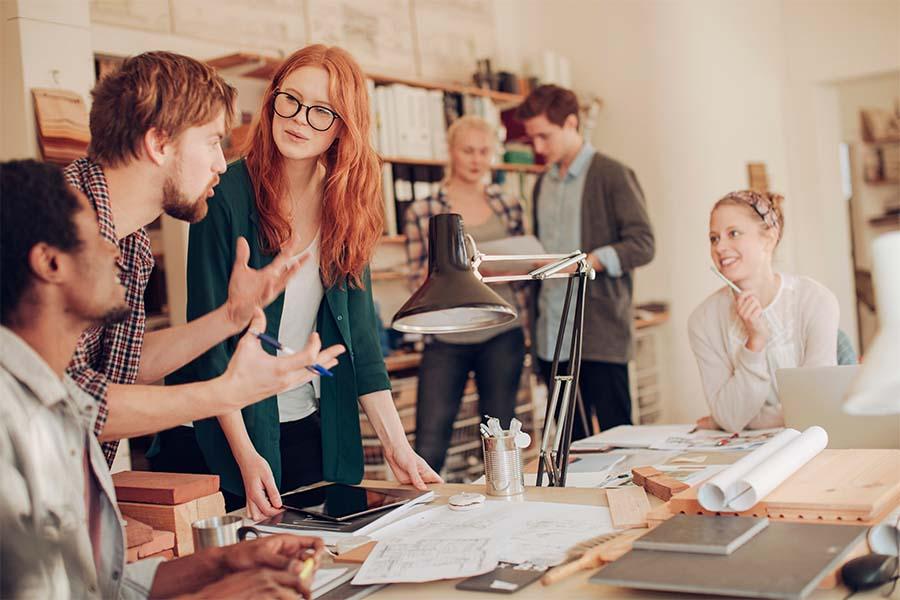 A collaborative office setting with people engaged in discussion and work around a large table covered with papers, blueprints, and office supplies, highlighting teamwork and creative brainstorming.