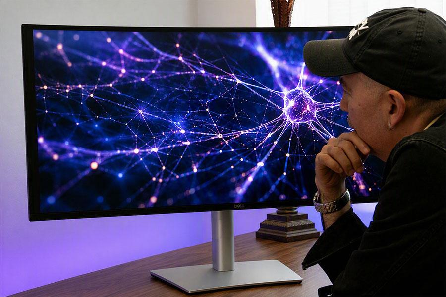Photograph showing a person viewing a computer monitor displaying an abstract neural-network visualization. Purple and blue glowing nodes connected by white lines form a dense web.