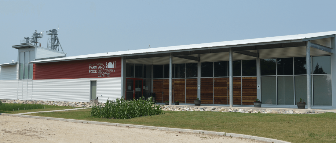 The front of the Bruce D. Campbell Farm and Food Discovery Centre. The sky behind the building is grey and cloudy, the grass in front of the building is lush and green.
