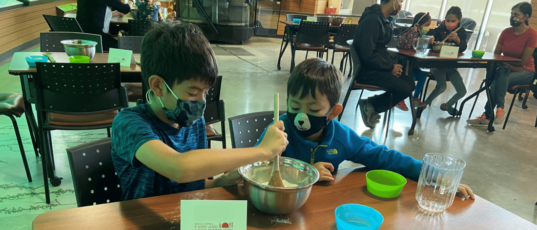 Two children sit at a wooden table wearing masks. One is mixing pizza dough in a metal bowl with a wooden spoon while the other watches. In the background a family of four sits at another wooden table where one child is mixing their dough and the other child and two parents watch. 