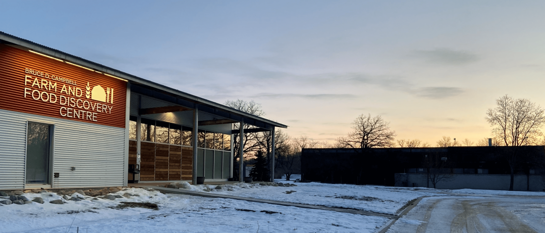 The sun is rising just east of the Bruce D. Campbell Farm and Food Discovery Centre. There is a light dusting on snow on the ground and the building's logo is lit up.