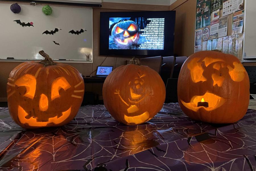 Three jack-o-lanterns that were carved at FFDC are glowing in a dark room. In the background there are halloween decorations and a youtube playlist displayed on the TV.