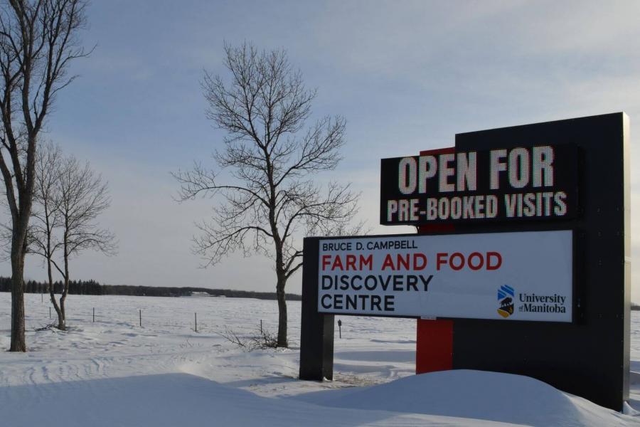 A digital sign at the entrance of the Farm and Food Discovery Centre indicates the facility is open for pre-booked visits. The sign is surrounded by mounds of white snow and bare trees on a winter morning. 