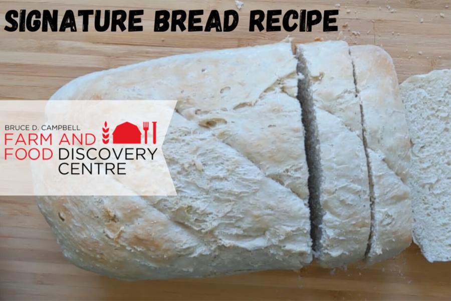 A partially slices loaf of homemade bread sits on top of a wooden cutting board titles "Signature Bread Recipe".