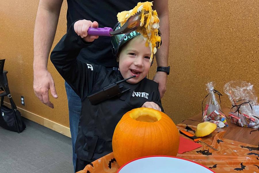 A young child smiles gleefully while pulling out the guts of a pumpkin while carving a jack-o-lantern. The child is wearing a swat costume while their parent stands behind watching.