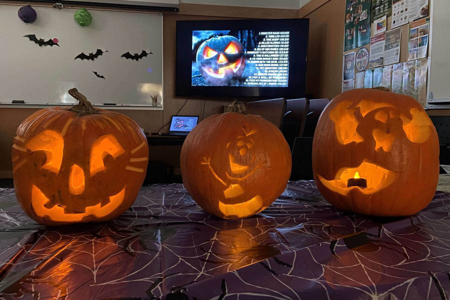 Three jack-o-lanterns that were carved at FFDC are glowing in a dark room. In the background there are halloween decorations and a youtube playlist displayed on the TV. 