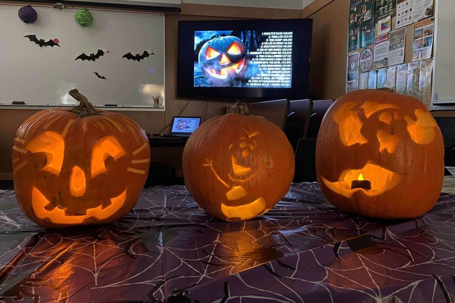 Three jack-o-lanterns that were carved at FFDC are glowing in a dark room. In the background there are halloween decorations and a youtube playlist displayed on the TV.