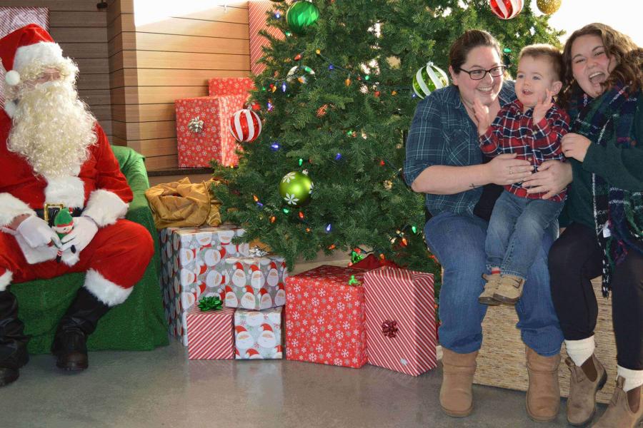 Two adults smile gleefully at a young child between them as they pose for photos with Santa at FFDC. The guests are sitting on a hay bale to the right of a large decorated tree with wrapped gifts underneath. On the left of the tree, Santa sits on a green chair looking at the child with a smile. 