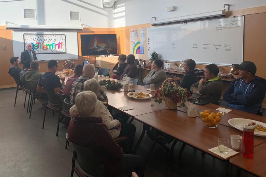 A group of 20 adults rents the FFDC classroom for a combination holiday and retirement party. They are sitting around a long table chatting with retirement and winter holiday decorations around the room.