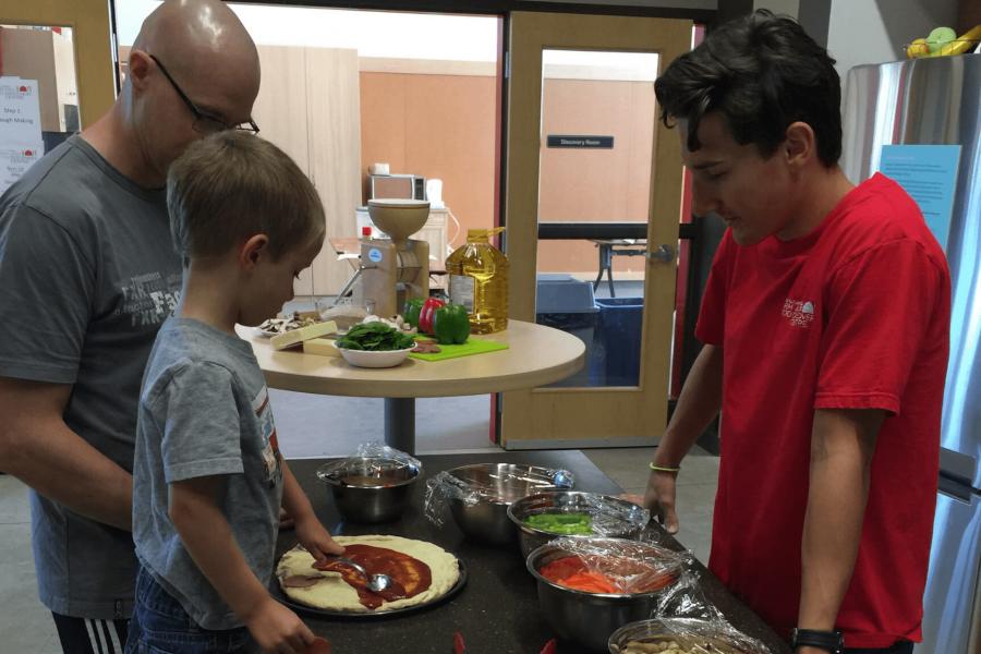 A volunteer helps a young child and adult top their pizza during the Pizza Party family fun event.