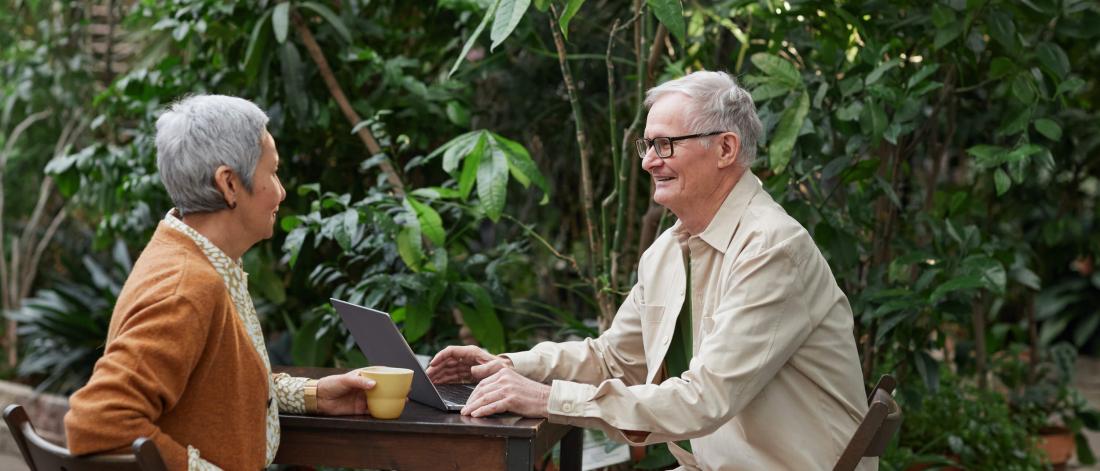 A middle aged couple sit at a table having coffee with a laptop open while they talk. The background is full of lush greenery. 