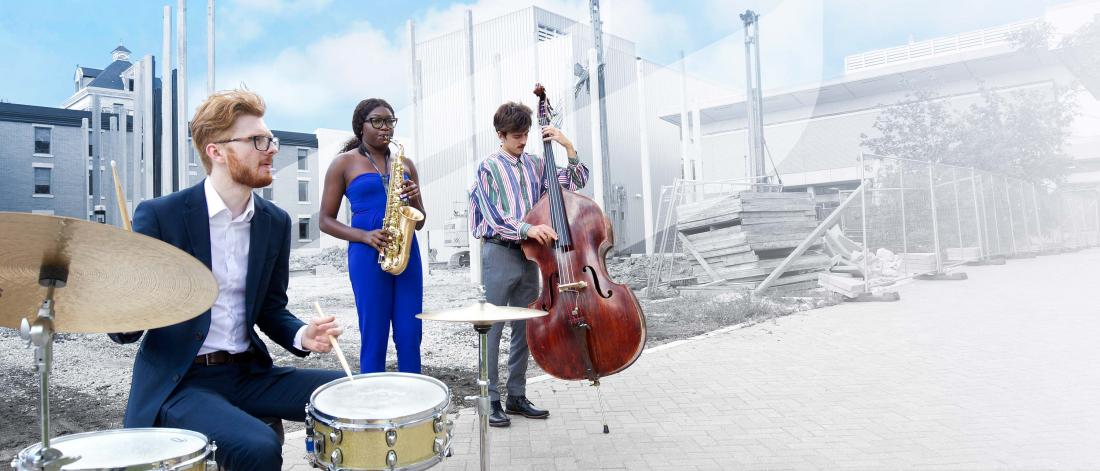 A trio of students playing drums, saxophone and bass on the sidewalk in front of the construction site for the new concert hall