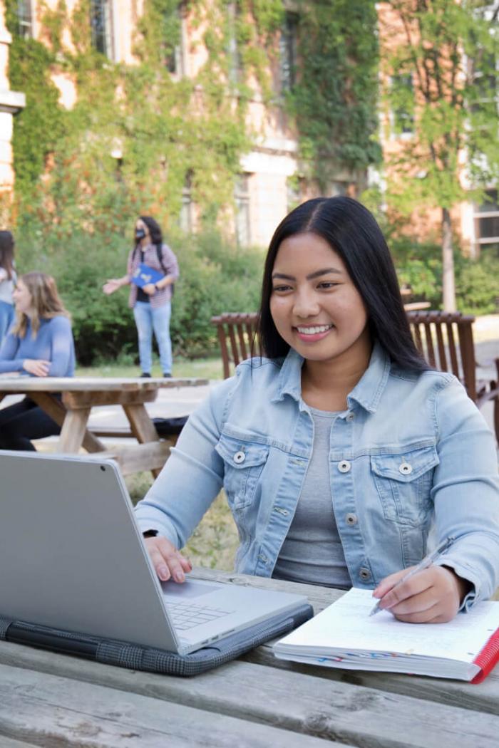 A student takes notes while studying at her laptop outdoors seated at a picnic table. 