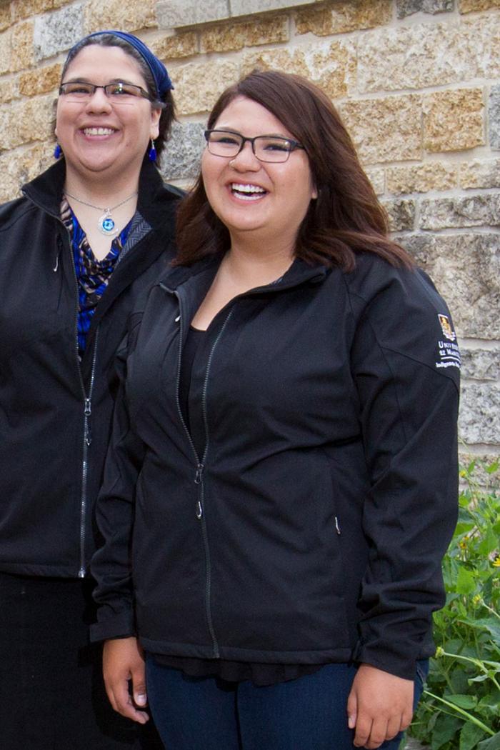 Selena Suderman and Chelsey Meade, two young Indigenous women standing outside of Migizii Agamik Bald Eagle Lodge a modern limestone clad building