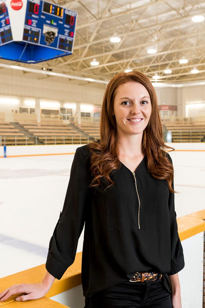 Caitlyn Fyten a smiling young Caucasian woman with red hair and freckles stands in front of the hockey ring boards with the rink and stands in the background.