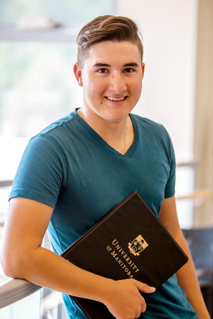Riley Chartrand, a smiling young Metis male stands leaning against a railing with a University of Manitoba clipboard in hand in a light and airy space.