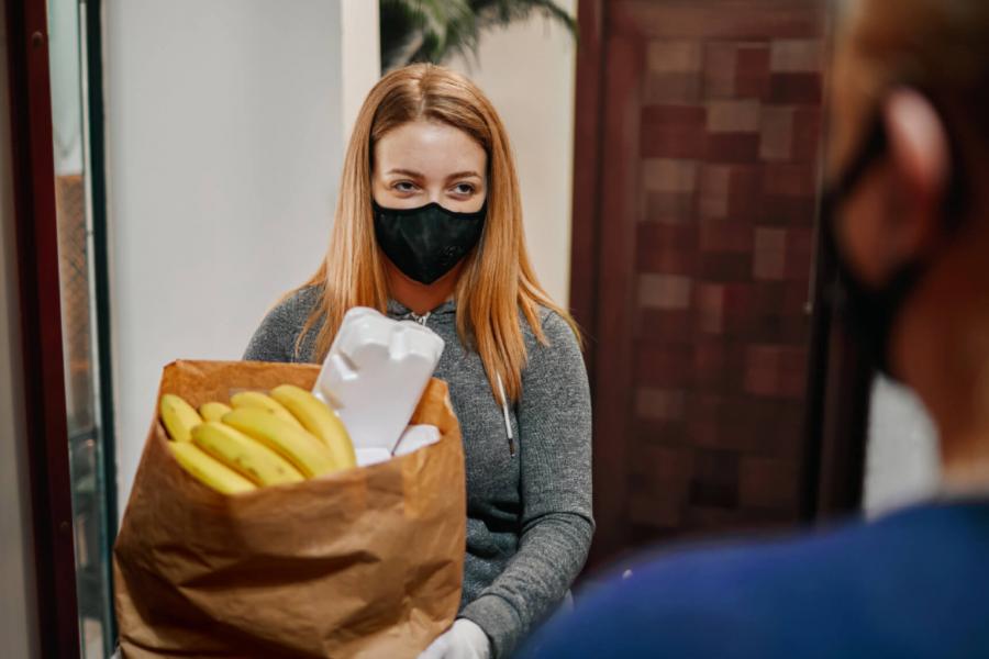 A student wearing a mask and gloves holds a grocery bag full of food.