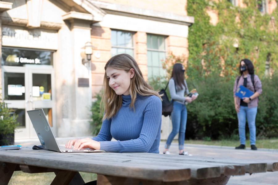 A student seated at a picnic table outdoors types on her laptop. Two other students can be seen in the background talking to each other. 